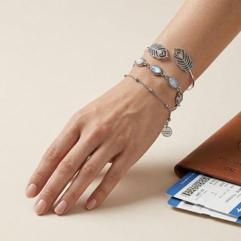 Hand wearing stacked silver bracelets: moonstone link bracelet, delicate bead chain and ornate feather cuff beside passport and boarding pass