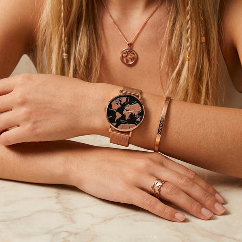 Model wearing a rose-gold world-map wristwatch with a glossy black mirrored dial, paired with a rose-gold Woodstock Zambon engraved cuff bracelet, matching world-map pendant necklace and ring — waterproof travel jewelry set in warm rose gold.
