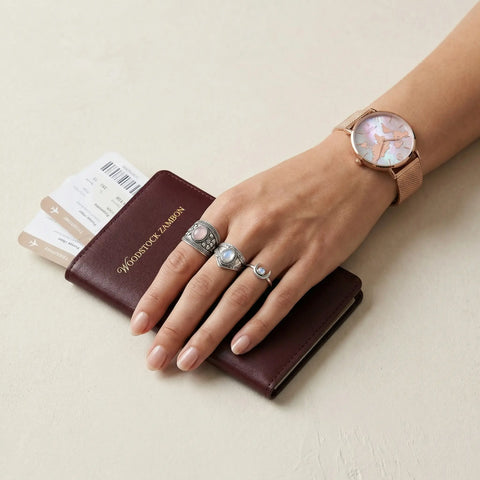 Hand wearing three ornate sterling silver moonstone rings beside a rose-gold mother-of-pearl world-map watch and passport — vintage boho travel jewelry