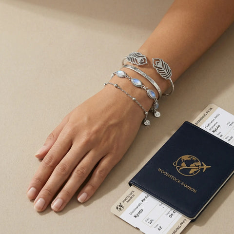 Hand wearing stacked silver bracelets: iridescent moonstone link bracelet, delicate bead chain and ornate feather cuff beside passport and boarding pass — waterproof travel-ready jewelry