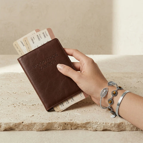 Close-up of a hand wearing Woodstock Zambon stacked silver bracelets: tiger-eye beaded bracelet, textured feather cuff and sleek herringbone cuff, styled with passport and boarding pass — waterproof travel-ready bracelets
